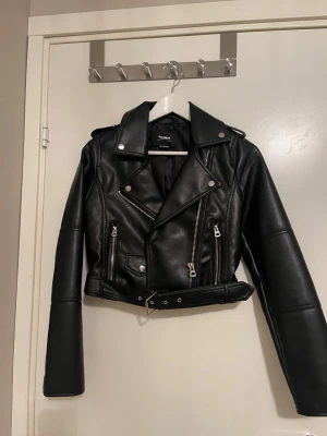 Svart croppad skinnjacka från NA-KD - ✨Snygg svart skinnjacka med croppad passform, klassisk bikerstil, dragkedjor, axelklaffar och bälte i midjan. Jackan har långa ärmar och coola detaljer som ger en edgy vibe till din outfit ✨