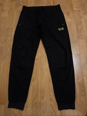 Svarta mjukisbyxor från EA7 - Svarta mjukisbyxor från EA7 med gul logga på vänster sida framtill. Byxorna har resår och snörning i midjan samt muddar vid bensluten. Två fickor med dragkedja framtill och dekorativa gula ränder baktill. Perfekta för en avslappnad stil.