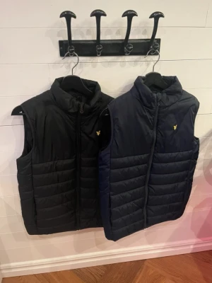 Svart och marinblå dunväst Lyle & Scott - Två stilrena dunvästar från Lyle & Scott, en i svart och en i marinblått. Båda har ståkrage, dragkedja framtill och den klassiska gula loggan på bröstet. Perfekt för lager-på-lager och snygg till både jeans och hoodie. Storlek 14-15 år på båda / hör gärna av er vid frågor ! 