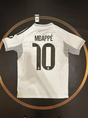 Real Madrid matchtröja Mbappé 10 - Vit Real Madrid matchtröja med Mbappé och nummer 10 på ryggen. Tröjan har svarta detaljer, klubbmärke på bröstet och sponsortryck framtill. Tillverkad i lätt och ventilerande material, perfekt för fotbollsträning eller match.