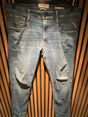 Replay Anbass ljusblå jeans - Snygga ljusblå Replay Anbass jeans med klassisk femficksdesign och diskreta slitningar. Jeansen har smal passform och är tillverkade i mjuk bomullsdenim med stretch för extra komfort. Perfekta för en avslappnad och trendig look.