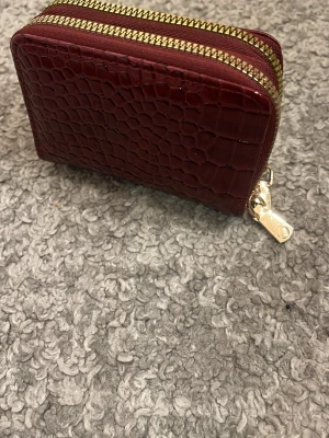 Vinröd plånbok med krokoprägling - Snygg vinröd plånbok med krokodilpräglat mönster och dubbla guldfärgade dragkedjor. Plånboken är kompakt och har en lyxig känsla tack vare det glansiga skinnmaterialet. Perfekt för kort och mynt.