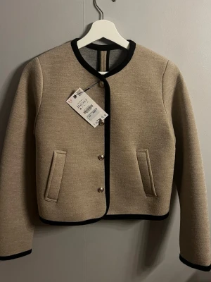 Beige cardigan jacka med svarta detaljer - Stilren beige cardigan jacka från Zara med svarta kanter och guldfärgade knappar. Jackan har två fickor framtill och rund halsringning. Perfekt för dig som gillar en klassisk look med modern twist.