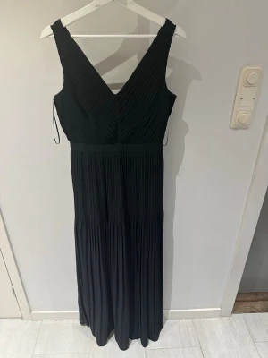 Svart långklänning från Esprit - Elegant svart långklänning från Esprit med v-ringad omlott-topp och plisserad kjol. Klänningen är ärmlös och har en stilren, tidlös design som passar perfekt till speciella tillfällen. Tillverkad i ett mjukt och följsamt material.