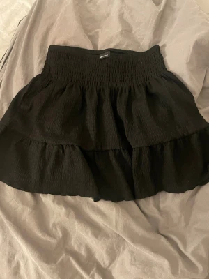 Svart volangkjol från Gina Tricot - Supersöt svart kortkjol från Gina Tricot med bred smock i midjan och dubbla volanger. Kjolen har en flowy passform och är perfekt för att skapa en trendig och avslappnad look. Tillverkad i mjukt tyg som känns skönt mot huden.