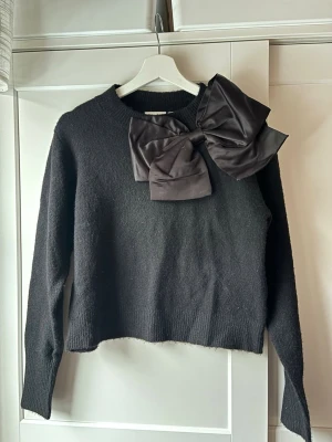 Stickad tröja med stor rosett - Säljer en svart stickad tröja från Kappahl med en stor rosett framtill som ger plagget en elegant och unik touch. Perfekt för dig som vill ha något som sticker ut men ändå är stilrent. Den är i storlek xs