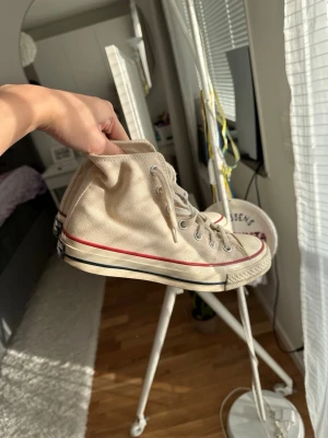 Beige höga Converse sneakers med röd rand - Säljer ett par Converse klassiska beige höga sneakers med vit sula och röd rand runt sulan. Skorna har snörning och metallhål för skosnören. Tidlös design som passar till det mesta.