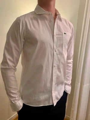 Vit skjorta från Lacoste - Klassisk vit skjorta från Lacoste i nyskick! Skjortan har en bröstficka med den ikoniska gröna krokodilloggan broderad. Tillverkad i bomull och har en normal passform. Perfekt för en clean och stilren look. Storlek 176cm/16
