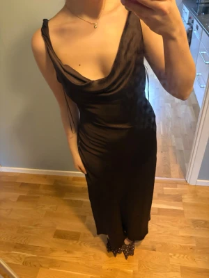 Brun långklänning med draperad urringning - Säljer en elegant brun långklänning med draperad urringning och smala axelband. Klänningen har en figurnära passform och är tillverkad i ett glansigt material som ger en lyxig känsla. Perfekt för dig som vill ha en stilren och trendig look.