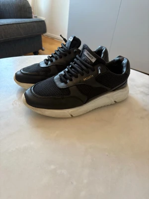 Svarta sneakers från Axel Arigato - Stilrena svarta sneakers från Axel Arigato med vit sula och detaljer i mesh och skinn. Skorna har snörning och diskret logga i guld på sidan. Perfekta för dig som gillar en modern och sportig look.
