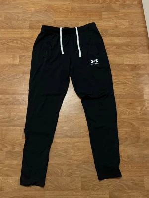 Svarta mjukisbyxor från Under Armour - Svarta mjukisbyxor från Under Armour med vit dragsko i midjan och logga på vänster lår. Byxorna har smal passform och resår i midjan. Perfekta för chill eller träning.