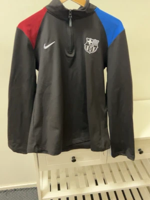 Svart Nike FC Barcelona tracksuit - Svart långärmad träningströja från Nike med FC Barcelona-logga på bröstet. Tröjan har dragkedja vid halsen och färgblock i rött och blått på axlarna. Perfekt för fotbollsträning eller sportiga aktiviteter.
