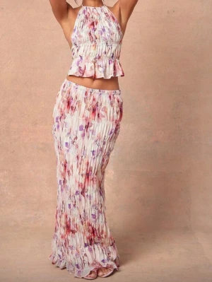 Blommigt tvådelat set med volanger - Säljer ett somrigt tvådelat set med blommönster i rosa, lila och rött på vit botten. Toppen är ärmlös med halterneck och volangkant, kjolen är lång och plisserad med elastisk midja. Perfekt för dig som gillar boho-stil och lätta material.