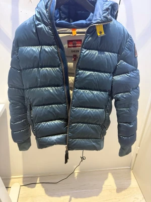 Parajumpers puffer - Snygg blå pufferjacka från Parajumpers med huva och dragkedja framtill. Jackan har quiltad design och ribbade muddar vid ärmslut och nederkant. Perfekt för kyliga dagar och har en modern, sportig look.