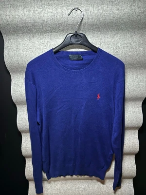 Blå tröja från Polo Ralph Lauren - Klassisk blå rundhalsad tröja från Polo Ralph Lauren med den ikoniska röda logotypen broderad på bröstet. Tunn och mjuk kvalitet, perfekt för lager på lager. Långa ärmar och ribbade muddar vid ärmslut och nederkant.