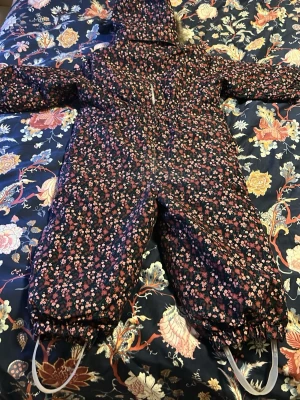 Blommig overall från Name It - Mörk overall från Name It med blommigt mönster i rosa och rött. Vattenoch vindtät overall med huva, dragkedja framtill och elastiska band vid bensluten. Perfekta för kalla dagar och vinteraktiviteter.