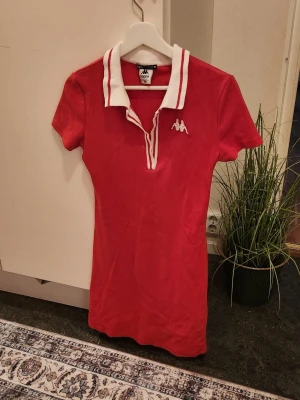 Röd kort klänning från Kappa - Sportig röd klänning från Kappa med vit krage och vita detaljer längs knäppningen. Klänningen har korta ärmar, figurnära passform och klassisk Kappa-logga på bröstet. Perfekt för dig som gillar retrostil och vill sticka ut.