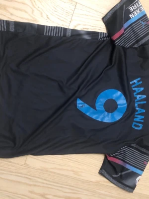Manchester City Haaland tröja svart - Säljer en svart Manchester City fotbollströja från Puma med Haaland och nummer 9 på ryggen. Tröjan har blå detaljer, klubbmärke och sponsorloggor framtill samt mönstrade ränder. Perfekt för fotbollsfans!