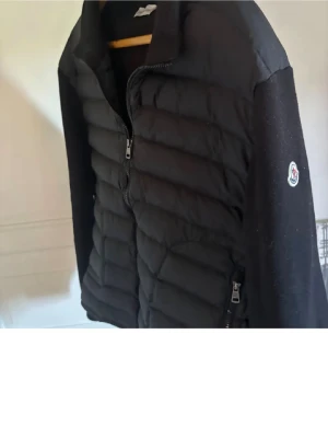Moncler Cardigan - Svart Cardigan från Moncler med quiltad front och släta ärmar. Jackan har dragkedja framtill och Moncler-logga på ärmen. Perfekt för kyliga dagar och ger en clean look.