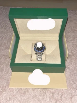 Stilren silverfärgad klocka i box - Säljer en snygg klocka med silverfärgad länkarmband och rund urtavla. Klockan har en svart och blå vridbar bezel med tydliga siffror och detaljer. Kommer i en exklusiv grön och beige box som ger en lyxig känsla. Perfekt accessoar för dig som gillar klassisk design.