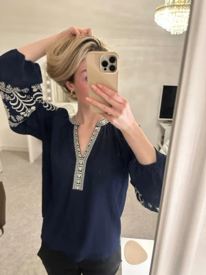 Mörkblå blus med broderi - Snygg mörkblå blus med vita broderade blommor på ärmarna och dekorativt broderi längs v-ringningen. Blusen har vida, långa ärmar och är tillverkad i ett lätt, luftigt tyg.