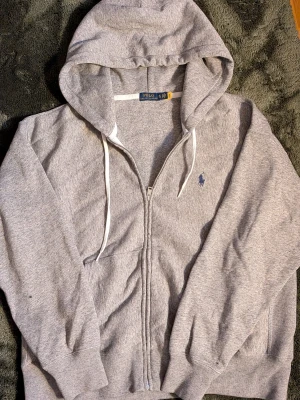 Grå zip hoodie från Polo Ralph Lauren - Klassisk grå hoodie med dragkedja från Polo Ralph Lauren. Har huva med vita snören, två fickor fram och den ikoniska blå loggan broderad på bröstet. Tillverkad i mjuk bomullsblandning för maximal komfort och stil. (Obs kan diskutera pris) 