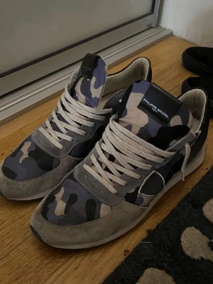 Philippe Model sneakers med camomönster - Snygga sneakers från Philippe Model med låda medföljer. blått och grått camouflagemönster, vita snören och detaljer i mocka. Skorna har en klassisk rund tå och platt sula, perfekt för dig som gillar streetstyle och unika detaljer.