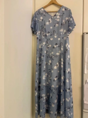 Blommig blå långklänning - Ljusblå långklänning med blommigt mönster och v-ringning. Klänningen har korta ärmar och dragkedja i ryggen. Perfekt för vår och sommar, med en luftig och bekväm passform.