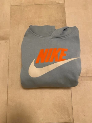 Blå Nike hoodie - Hej! Säljer en ljusblå hoodie från Nike med stor vit swoosh och orange Nike-tryck på bröstet. Klassisk ”känguruficka” framtill och huva. Perfekt för en avslappnad och sportig stil. Om det ser ut som missfärgning på tröjan är det inte det, bara min kameras fel😊.Hör gärna av dig vid frågor!