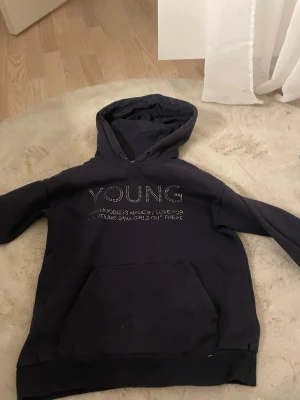Svart hoodie med texten YOUNG - Svart hoodie med huva och ficka framtill. På bröstet står det 'YOUNG' i glittriga stenar och under finns en mindre text. Hoodien har en klassisk passform och ribbade muddar vid ärmslut och nederkant. Perfekt för en chill och avslappnad stil.