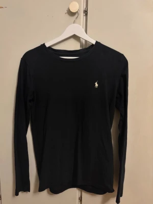 Svart långärmad topp Polo Ralph Lauren - Klassisk svart långärmad topp från Polo Ralph Lauren med rund halsringning och den ikoniska loggan broderad på bröstet. Tillverkad i mjuk bomull som känns skön mot huden. Perfekt basplagg till alla outfits.