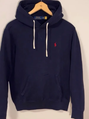 Mörkblå hoodie från Polo Ralph Lauren - Mörkblå hoodie från Polo Ralph Lauren med vit dragsko i huvan och röd broderad logga på bröstet. Klassisk känguruficka framtill och ribbade muddar vid ärmar och nederkant. Perfekt för en avslappnad och stilren look.