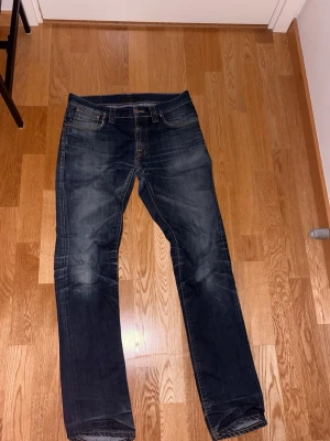 Mörkblå jeans från Nudie Jeans - Säljer ett par mörkblå jeans från Nudie Jeans med klassisk femficksdesign och snygga slitningar. Jeansen har en liten lagning se på bil. Skriv vid funderingar. 