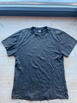 Mörkgrå t-shirt från Reclaimed Vintage strl M - En klassisk svart t-shirt från Reclaimed Vintage i mjuk bomull. T-shirten har rund halsringning och korta ärmar, perfekt för en enkel och avslappnad stil. Passformen är normal och tyget känns skönt mot huden.