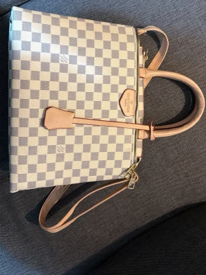Rutig handväska från Louis Vuitton - Säljer en snygg rutig handväska från Louis Vuitton i beige och vitt med klassiskt Damier-mönster. Väskan har både handtag och axelrem i ljust skinn samt guldfärgade metalldetaljer och dragkedja. Perfekt för dig som vill ha en ikonisk och stilren accessoar.