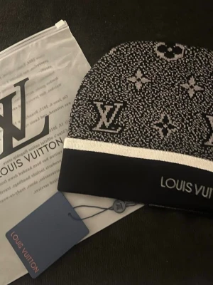 svartvit mössa - Snygg svartvit mössa med klassiskt monogrammönster och logotyp framtill. Mössan har en bred svart kant med vit rand och är tillverkad i mjukt stickat material. Perfekt accessoar för dig som gillar exklusiv streetstyle.