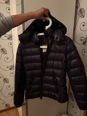 Svart Moncler pufferjacka med huva - Snygg svart pufferjacka från Moncler med huva och klassisk logga på ärmen. Jackan har quiltad design, dragkedja framtill och två fickor med dragkedja. Perfekt för dig som vill ha en stilren och varm jacka med exklusiv känsla. Klisterlappen (serien) har lossnat en gång så sitter fast med superlim temporärt! BYTES helst mot en fjällräven jacka i storlek xs!!❤️