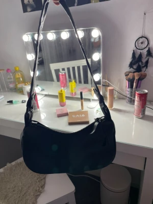 Svart axelväska i syntetmaterial - Säljer en stilren svart axelväska med croco-mönstrad yta på ena sidan och slät finish på den andra. Väskan har en justerbar axelrem och dragkedja upptill. Perfekt storlek för det viktigaste och passar till många olika looks.