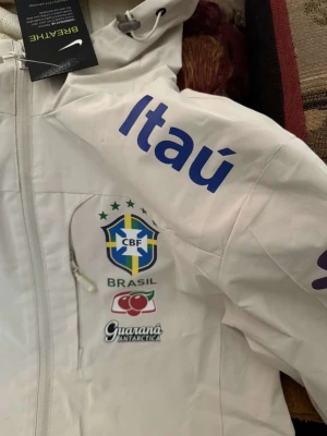Vit Nike Brasil vindjacka med huva - Vit vindjacka från Nike med Brasil CBF-logga, sponsormärken som Itaú och Guaraná Antarctica. Jackan har huva, hel dragkedja och sportig look. Perfekt för dig som gillar fotboll.