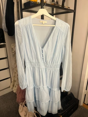 Ljusblå kortklänning från Maje - Supersöt ljusblå klänning från Maje med v-ringning, knappar framtill och långärmade ärmar. Klänningen har ett randigt mönster, volanger och smock i midjan för en skön passform. Perfekt för dig som gillar romantisk och feminin stil.