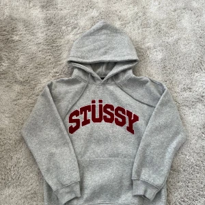 Grå dam Stüssy hoodie med röd logga - Säljer en grå hoodie från Stüssy i storlek M med stor röd Stüssy-text på bröstet. Tröjan har huva med snörning, ribbade muddar och klassisk känguruficka framtill. Perfekt för dig som gillar streetwear och vill ha en snygg och bekväm look.