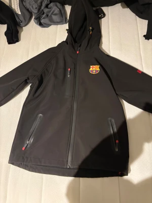 Svart vindjacka med FC Barcelona-logga - Svart vindjacka med huva och FC Barcelona-logga på bröstet. Jackan har hel dragkedja framtill, tre fickor med dragkedja och snygga röda detaljer på dragkedjorna. Perfekt för dig som vill visa ditt stöd för Barca och samtidigt hålla dig skyddad mot vinden.