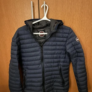 Mörkblå pufferjacka från Colmar - Säljer en riktigt snygg mörkblå pufferjacka från Colmar. Jackan har en dragkedja framtill och är quiltad för extra värme. Den har en huva och långa ärmar, perfekt för kyligare dagar.