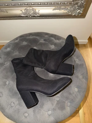 Svarta stövletter med klack - Säljer ett par svarta stövletter med hög klack och spetsig tå. Skorna har en stilren och smal siluett med platt sula och är tillverkade i ett syntetmaterial som ger en slät finish. Perfekta för dig som gillar minimalistisk och trendig stil. 8 cm