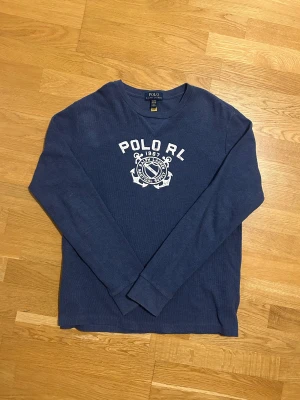 Blå långärmad tröja Polo Ralph Lauren - Snygg blå långärmad tröja från Polo Ralph Lauren i storlek jouniror XL vilken motsvarar herr medium. Tröjan har ett stort vitt tryck med logga och text på bröstet. Materialet är mjuk bomull med ribbade muddar vid ärmslut och hals. Perfekt för dig som gillar klassisk och sportig stil.