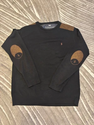 Svart stickad tröja Polo Ralph Lauren - Svart stickad tröja från Polo Ralph Lauren med bruna quiltade detaljer på axlar och armbågar. Klassisk rund hals och långärmad modell. Broderad logga på bröstet och armbågslapparna ger en snygg touch. Perfekt för dig som gillar stilrena och tidlösa plagg.