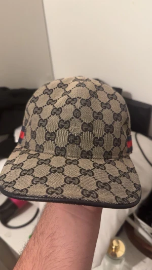 Gucci beige keps med GG-mönster - Säljer en beige Gucci keps med klassiskt GG-mönster i svart. Kepsen har en böjd skärm, justerbart spänne bak i svart skinn och röda/blå detaljer på sidorna. Materialet är bomull och polyester, perfekt för dig som vill ha en lyxig streetstyle-look.