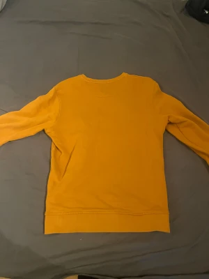 Gul sweatshirt från Lyle & Scott - Säljer en snygg gul sweatshirt från Lyle & Scott med klassisk rund hals och långärmad design. Tröjan har det ikoniska fågelmärket broderat på bröstet och ribbade muddar vid ärmslut och nederkant. Perfekt för dig som gillar stilrena och färgglada plagg.