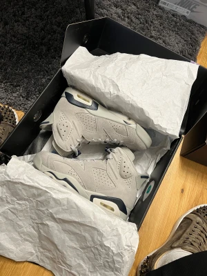 Nike Air Jordan 6 grå mocka sneakers - Nike Air Jordan 6 sneakers i ljusgrå mocka med marinblå detaljer och perforerade paneler. Snygg high-top siluett, snörning och synlig Air-enhet i sulan. Jumpman-logga broderad på hälen och platt sula med kontrastfärgade inslag. Perfekt för dig som gillar streetwear.