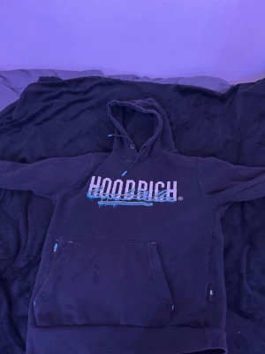 Svart hoodie från Hoodrich med blå detaljer - Svart hoodie från Hoodrich med stor vit och blå logga på bröstet och coolt grafiskt tryck. Tröjan har huva med snörning, känguruficka med dragkedja och blå detaljer på ärmen. Perfekt för dig som gillar streetwear och vill sticka ut.